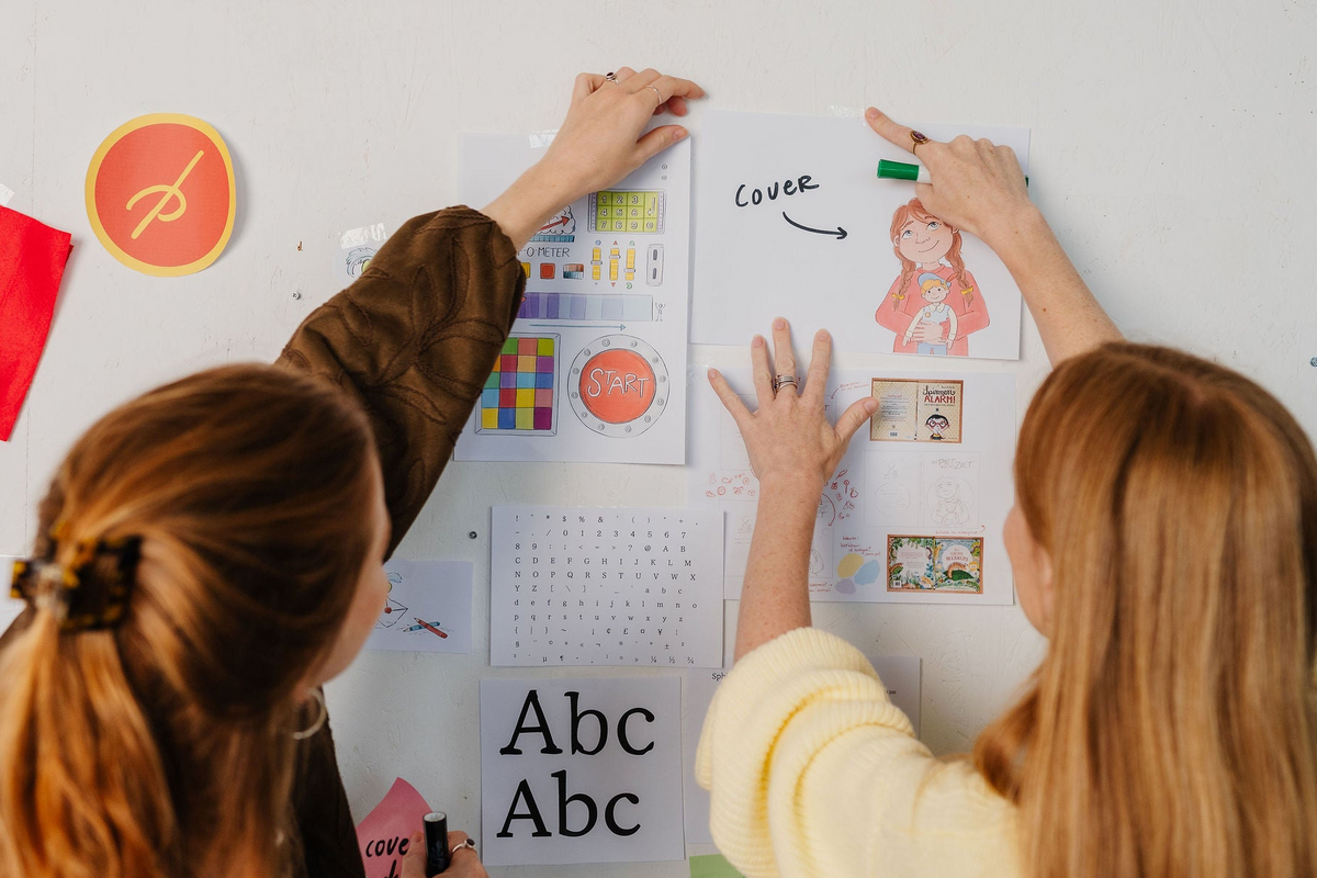 Twee makers hangen conceptillustraties en coverontwerpen van De Piet die 't Ziet op aan een moodboardmuur