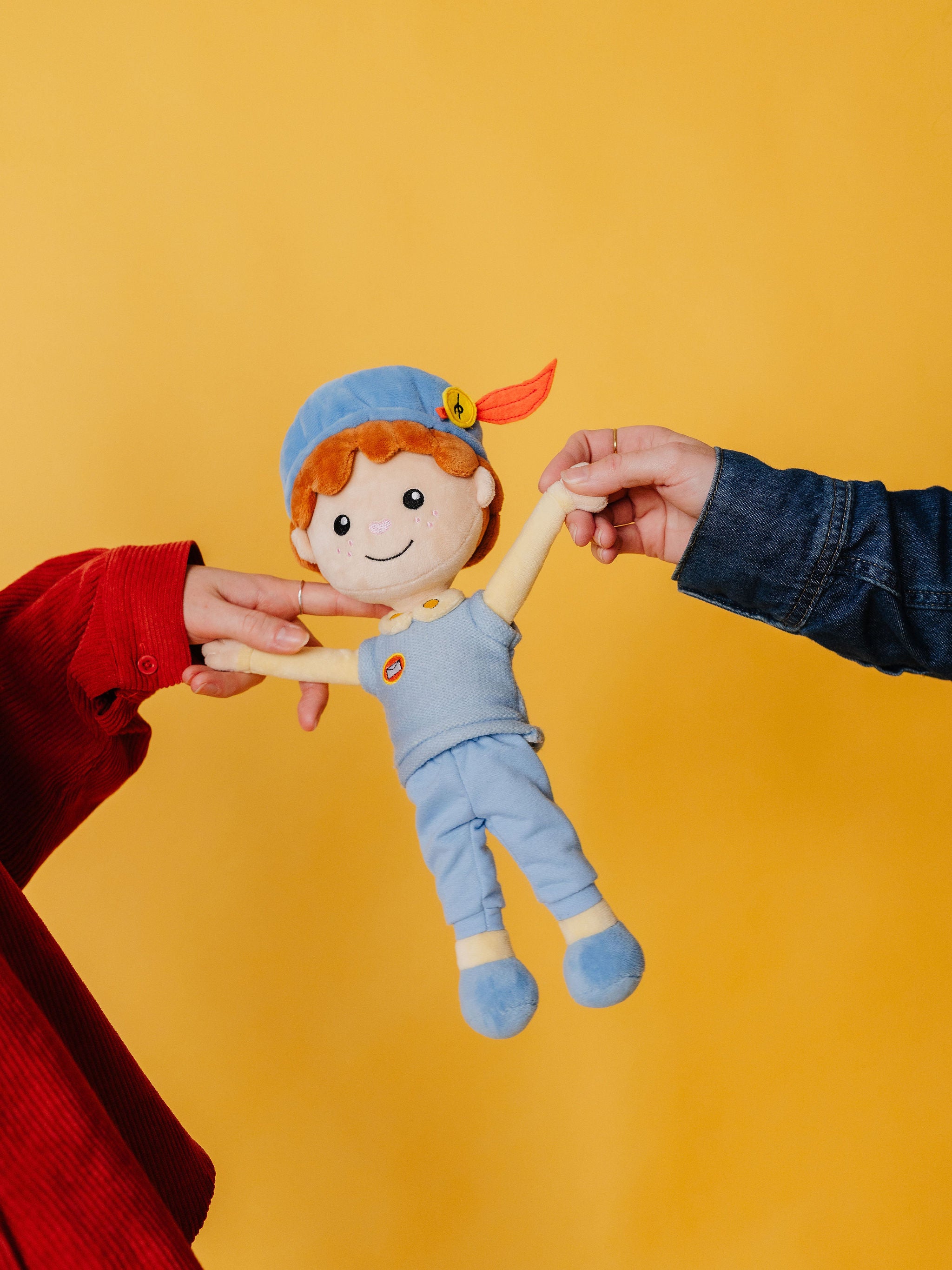 Twee handen bewegen de armen van de Piet die 't Ziet-knuffel tegen een gele achtergrond
