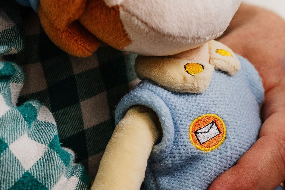 Close-up van het postembleem en de knopen op de overall van de Piet die 't Ziet-knuffel, vastgehouden door een kind in geruite pyjama