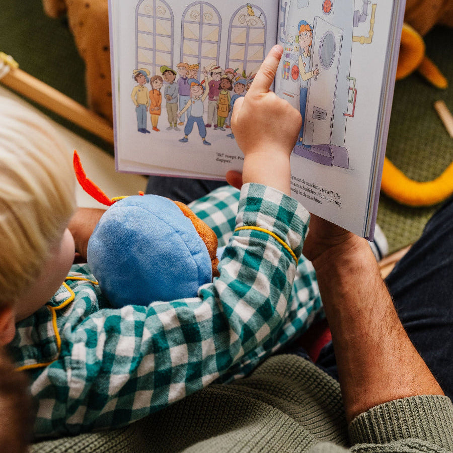 Kind wijst een pagina aan in het boek De Piet die 't Ziet terwijl een volwassene voorleest, met de knuffelpop ernaast