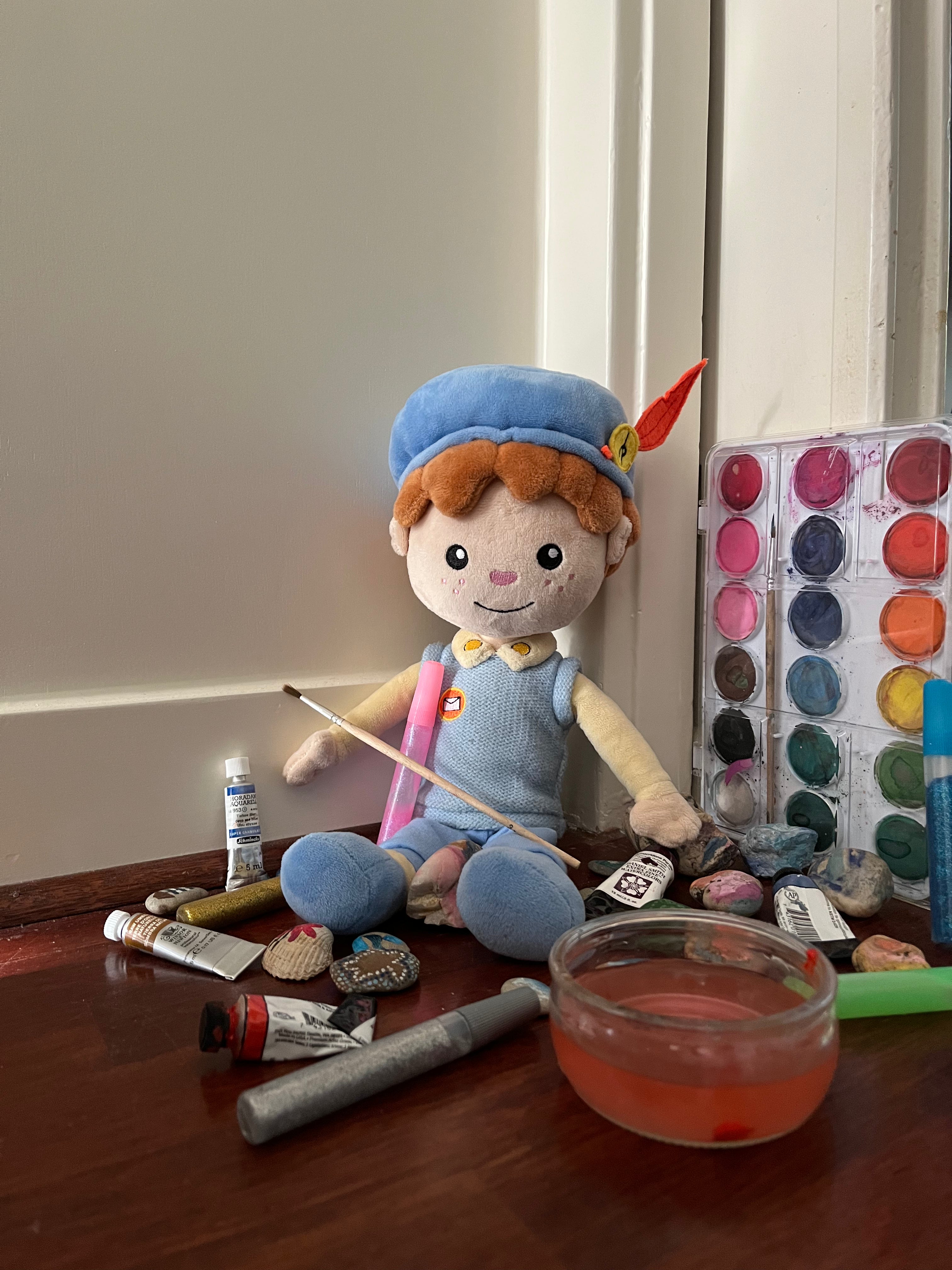 De Piet die 't Ziet-knuffel zit tussen verfspullen, waterverf en beschilderde stenen op een houten tafel