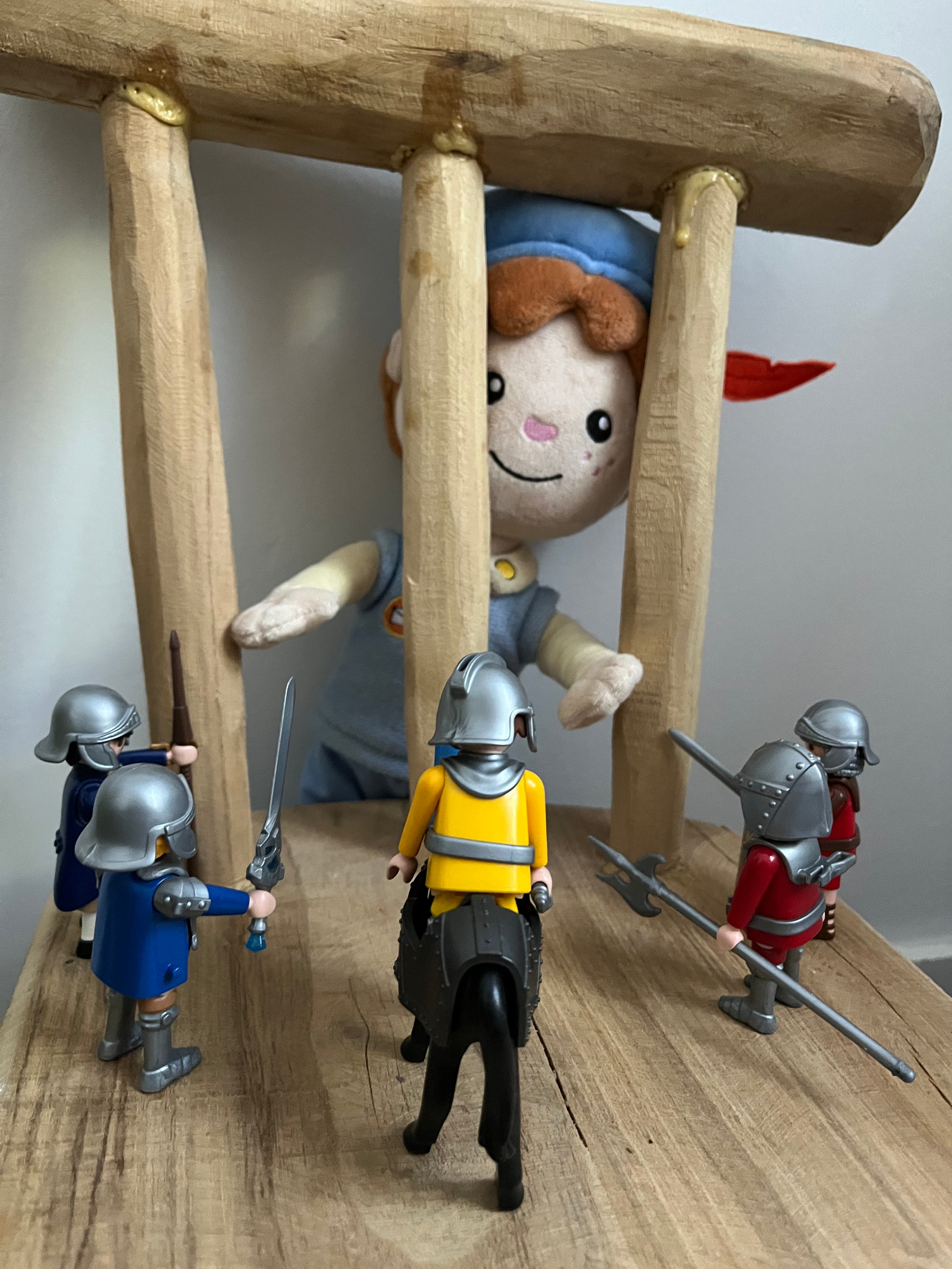 De Piet die 't Ziet-knuffel kijkt door de spijlen van een houten kasteelpoort naar een groep Playmobil-ridders